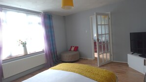 Une chambre à coucher avec un lit, une fenêtre avec des rideaux, une table de chevet, un téléviseur et une porte donnant sur une autre pièce.