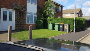 Une rue résidentielle bordée de maisons, avec une voiture garée et des bacs à déchets verts.