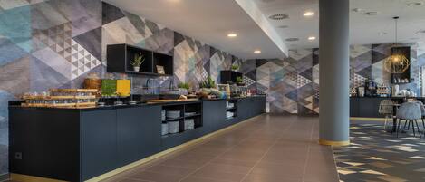 A modern hotel lobby with a long reception desk, a coffee station, and a seating area with chairs and a table.