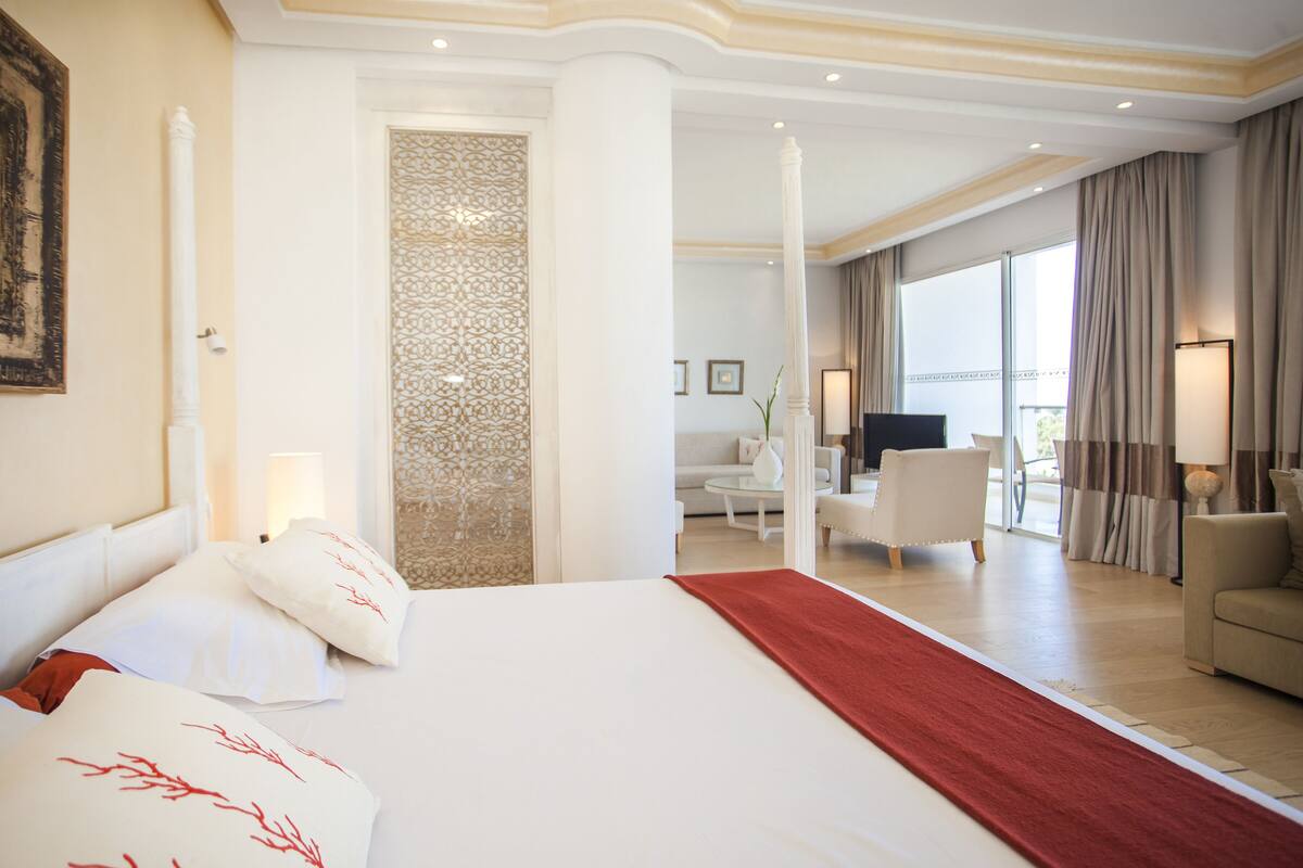 A hotel room with a large bed, a red runner, a sofa, and a TV in the background.