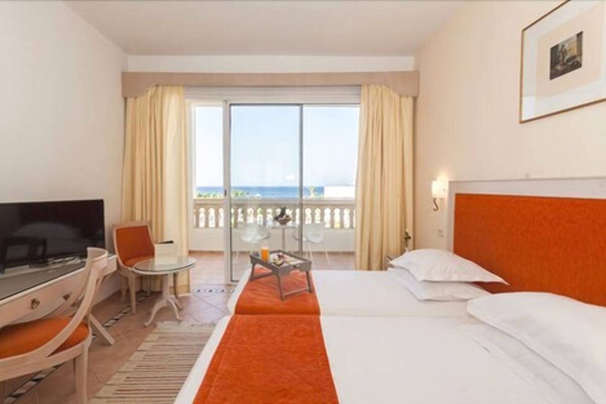 A hotel room with a bed, a desk with a TV, a small table with a drink, and a balcony with curtains.