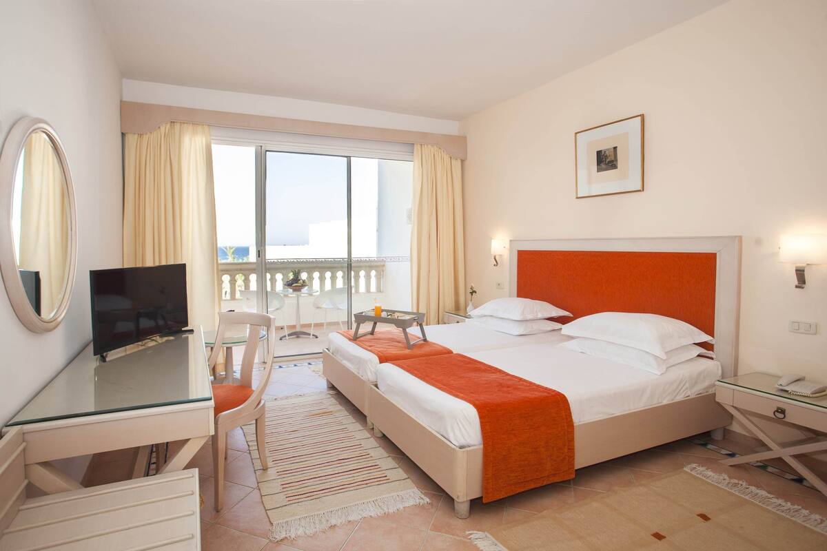 A hotel room with a bed, a desk with a TV, a chair, a balcony with curtains, and a window with a view.