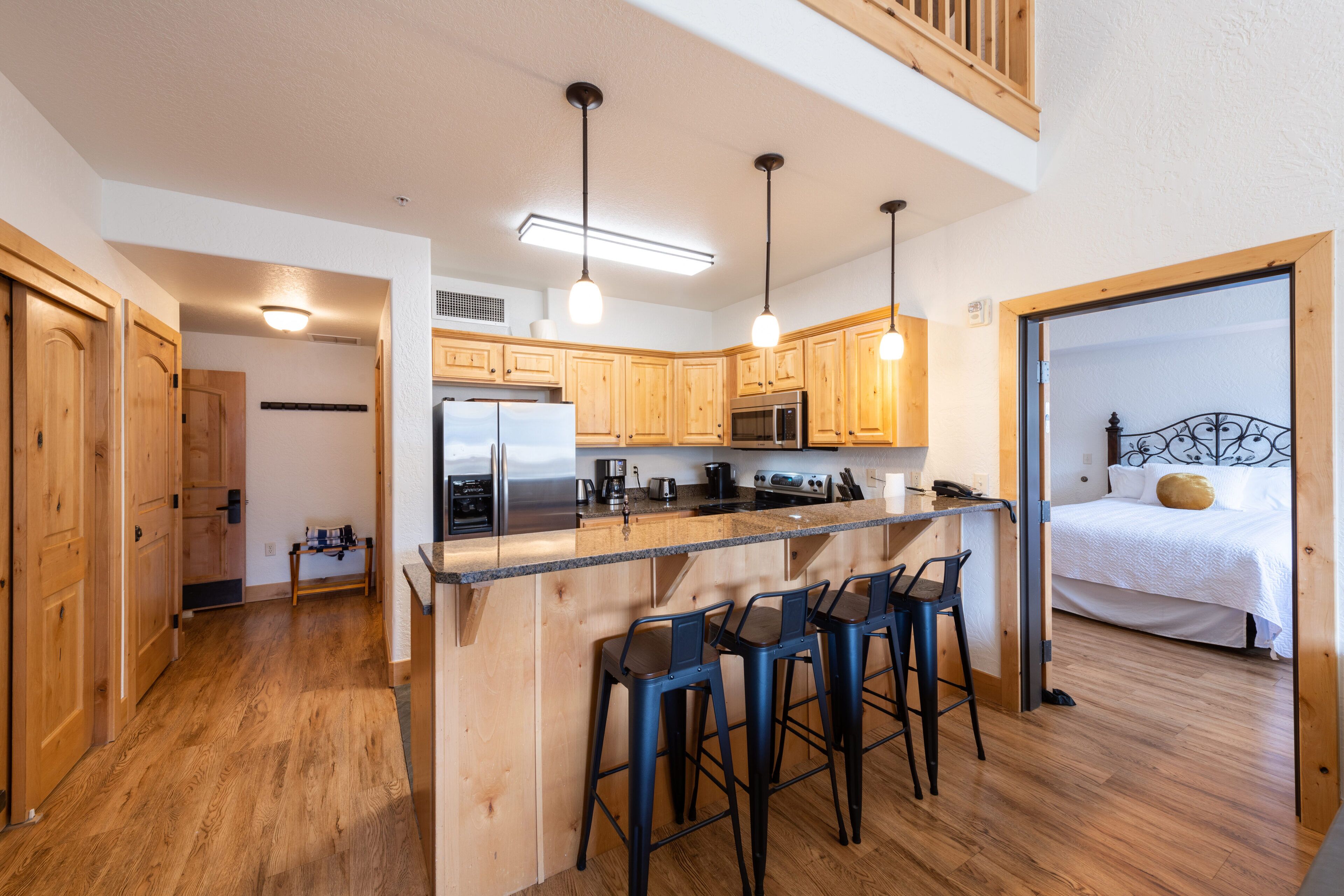A kitchen with wooden cabinets, a stainless steel refrigerator, a microwave, and a coffee maker. There are three bar stools and a wooden countertop.