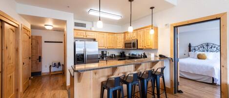 A kitchen with wooden cabinets, a stainless steel refrigerator, a microwave, and a coffee maker. There are three bar stools and a wooden countertop.