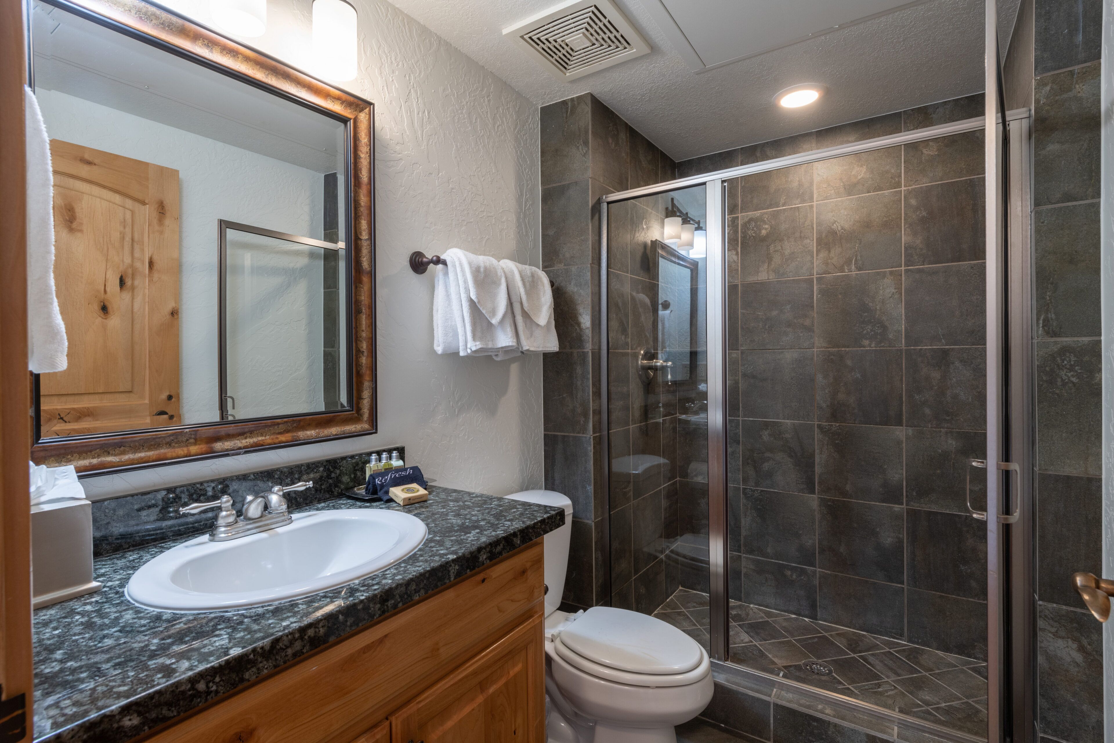 A bathroom with a granite countertop sink, wooden cabinets, a white toilet, and a glass-enclosed shower area.