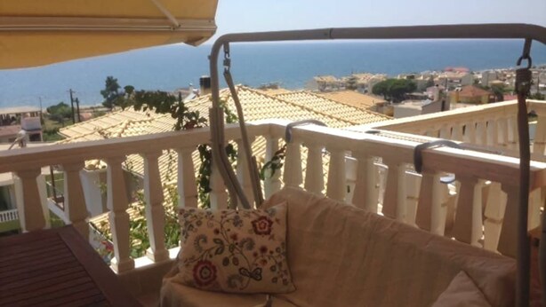 Un balcon avec un fauteuil suspendu, une table et une vue sur la mer et les bâtiments.