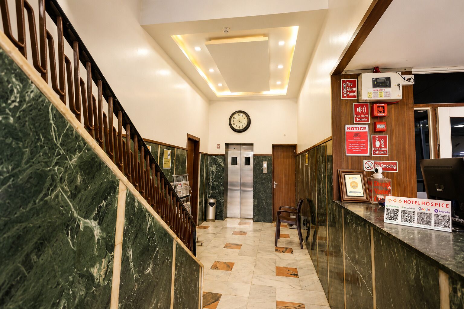 Un vestíbulo de hotel con una escalera de mármol, un mostrador de recepción, un reloj y un panel de alarma contra incendios.