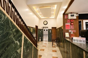 A hotel lobby with a marble staircase, a reception desk, a clock, and a fire alarm panel. - Hotel Hospice Near Surat Railway Station (Surat)