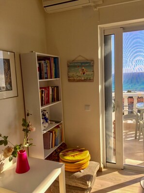A room with a bookshelf, a framed picture, a potted plant, and a view of the sea through a glass door.