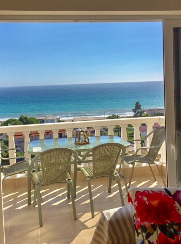 A balcony with a glass table and wicker chairs overlooking a beach and ocean.