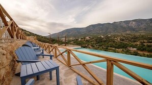 Una terraza de madera con sillas azules que da a una piscina con montañas al fondo.