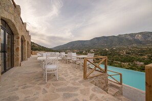 Un espacio al aire libre con piso de piedra y sillas blancas, con vista a una piscina turquesa y un paisaje montañoso.