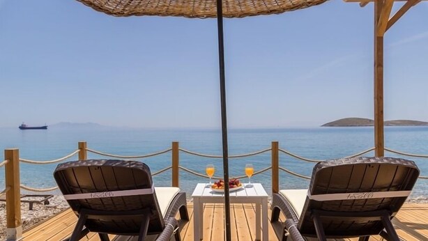 Una terraza de madera con dos sillones de mimbre orientados hacia un mar azul y despejado. En el centro, una mesa con una bandeja de frutas y dos vasos de zumo de naranja.