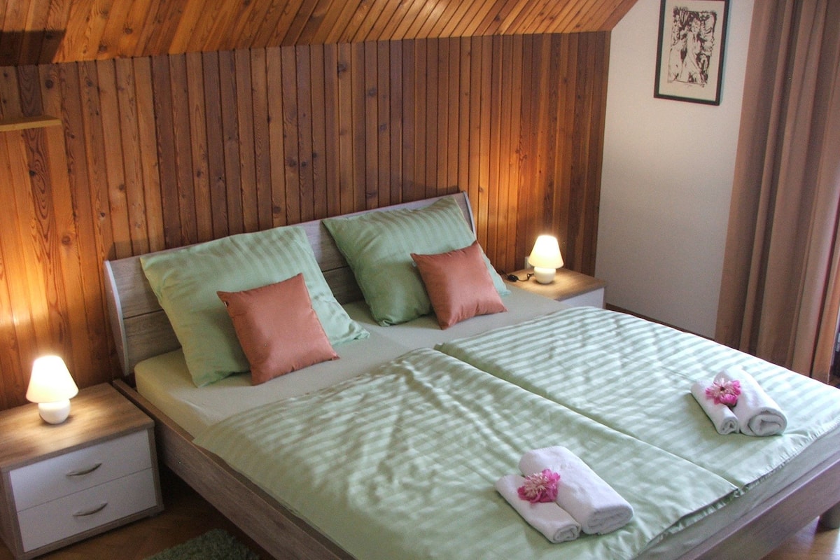 A neatly made bed with green bedding and pink pillows, two bedside lamps, and a small bedside table with a lamp.