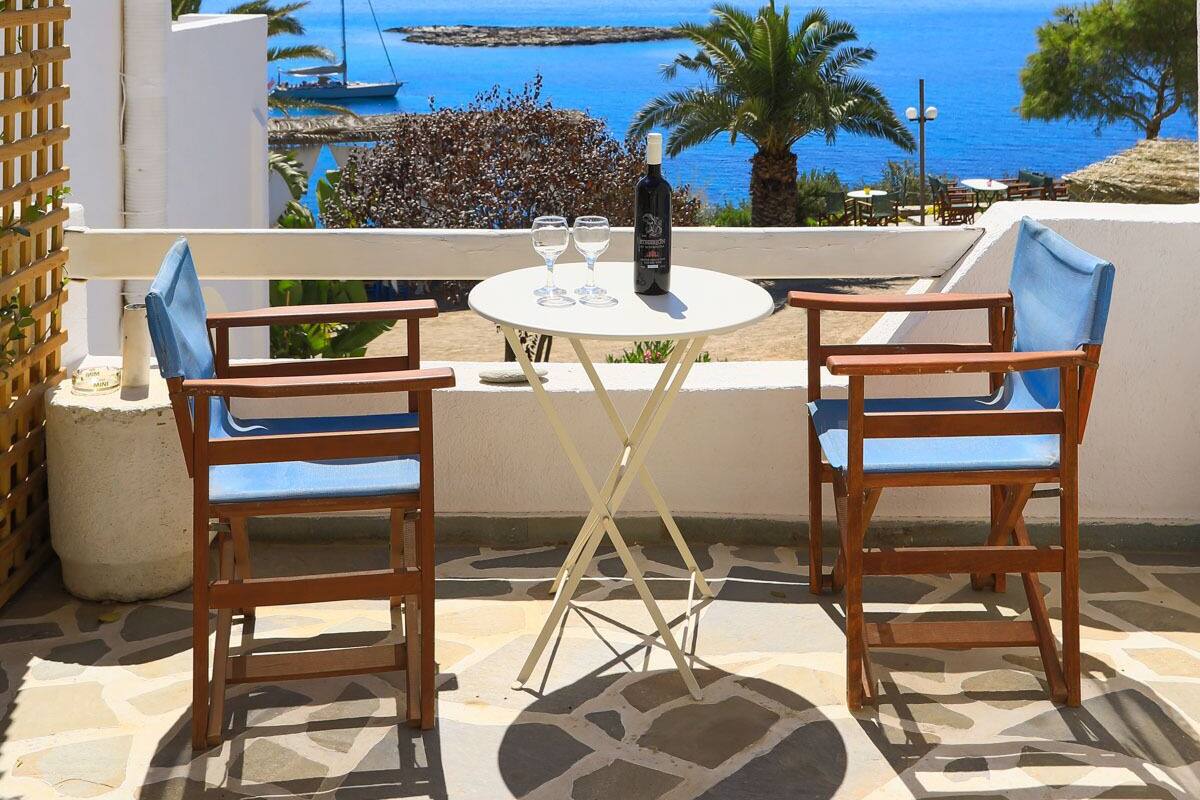 Une terrasse avec deux chaises en bois et une table, d’où l’on peut admirer une vue sur la côte, avec des bateaux et des palmiers.