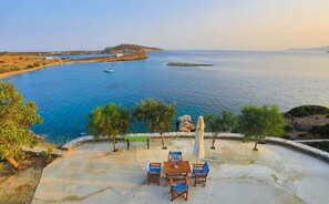Eine Terrasse mit Tisch und Stühlen mit Blick auf ein ruhiges Meer, wo Boote fahren, umgeben von viel Grün und felsigem Gelände.