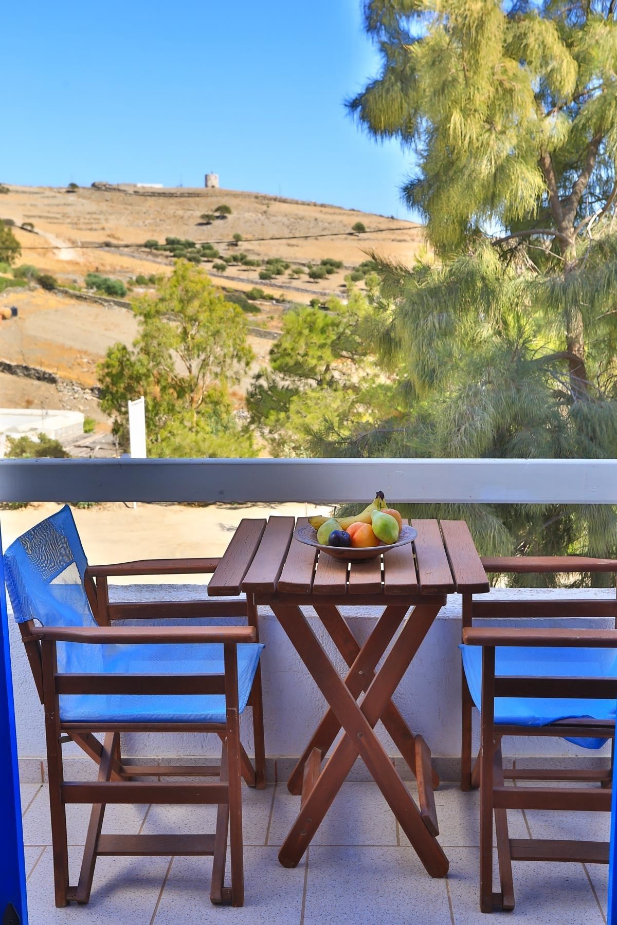 Una mesa de madera con un frutero, dos sillas azules y vistas a un paisaje montañoso con árboles.