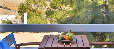 Una mesa de madera con un frutero, dos sillas azules y vistas a un paisaje montañoso con árboles.