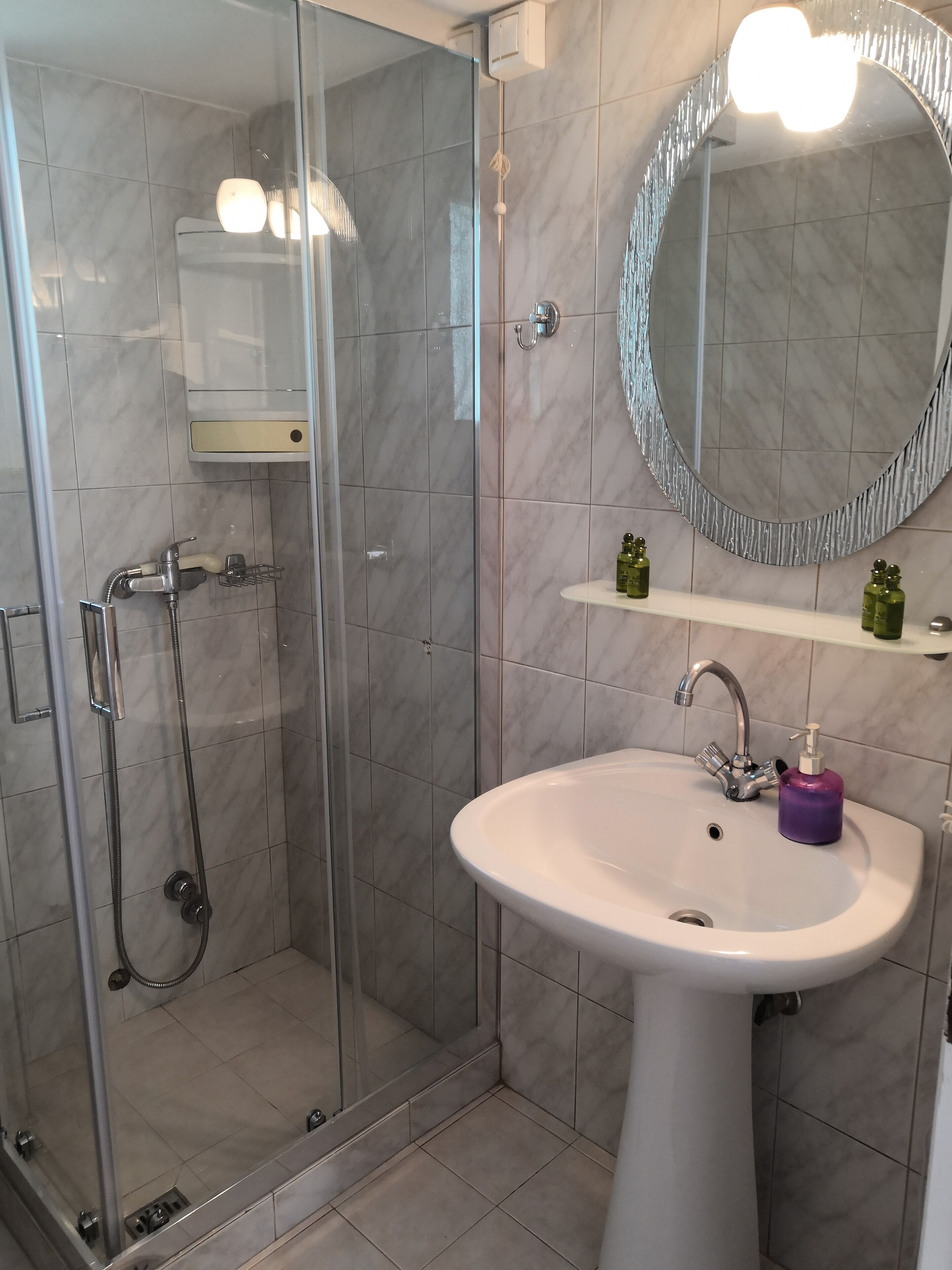 Un baño moderno con ducha acristalada, lavabo pedestal blanco, espejo redondo y una pequeña repisa con artículos de aseo.