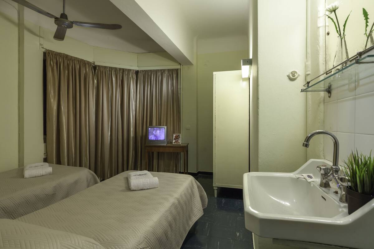 Une chambre d’hôtel avec deux lits, un ventilateur de plafond, un téléviseur posé sur une petite table, un lavabo avec un miroir et une plante en pot.