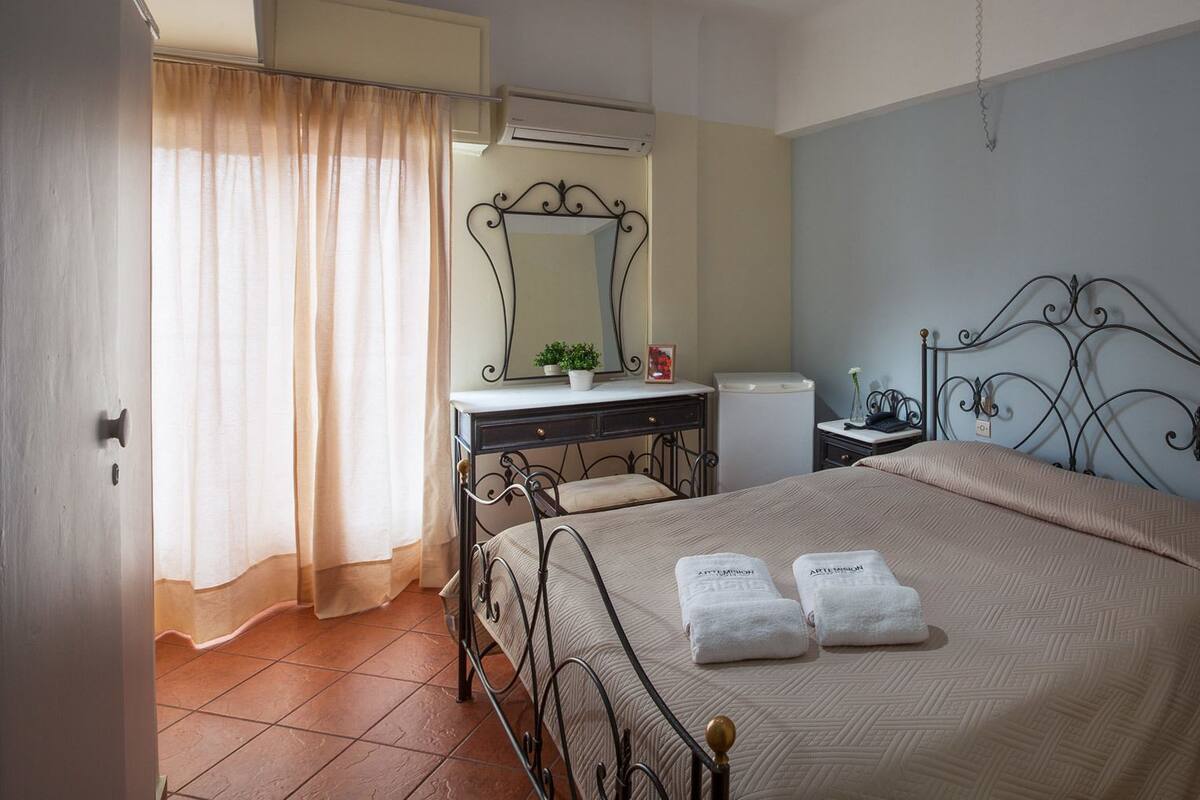 Une chambre à coucher comprenant un lit, une commode, une table de chevet, un miroir et une fenêtre avec des rideaux.