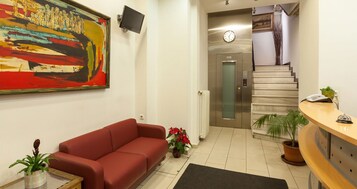 Egy előtér, melyben egy piros kanapé, egy nagyméretű, absztrakt festmény, egy óra és egy lépcsőház található, mely felvezet egy lifthez.