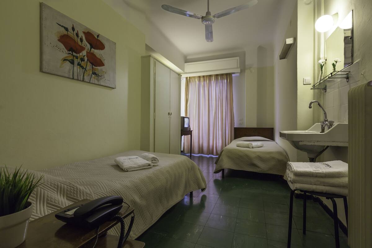 Une chambre d’hôtel avec deux lits, un ventilateur de plafond, une table de chevet avec un téléphone, une plante en pot et une salle de bain avec un lavabo et un miroir.