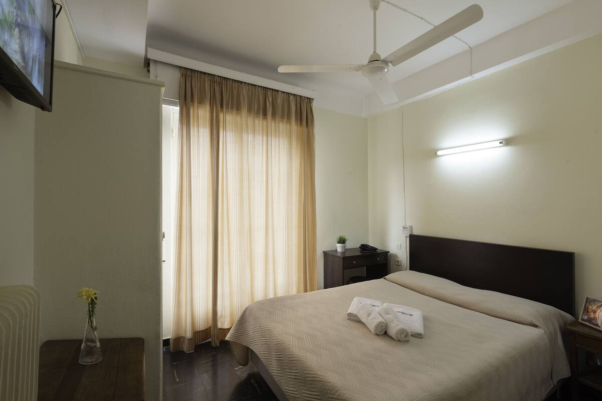 Une chambre d’hôtel avec un lit, une table de chevet ornée d’un vase, une commode, un ventilateur de plafond et une fenêtre avec des rideaux.
