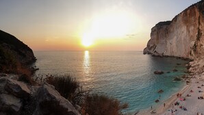 A coastal scene with a sunset, clear waters, rocky cliffs, and a beach with people. - Villa Eirini Ionian Home (Paxos)