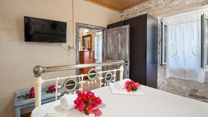 A neatly made bed with towels and flowers, a television on the wall, and a rustic stone wall. - Villa Eirini Ionian Home (Paxos)