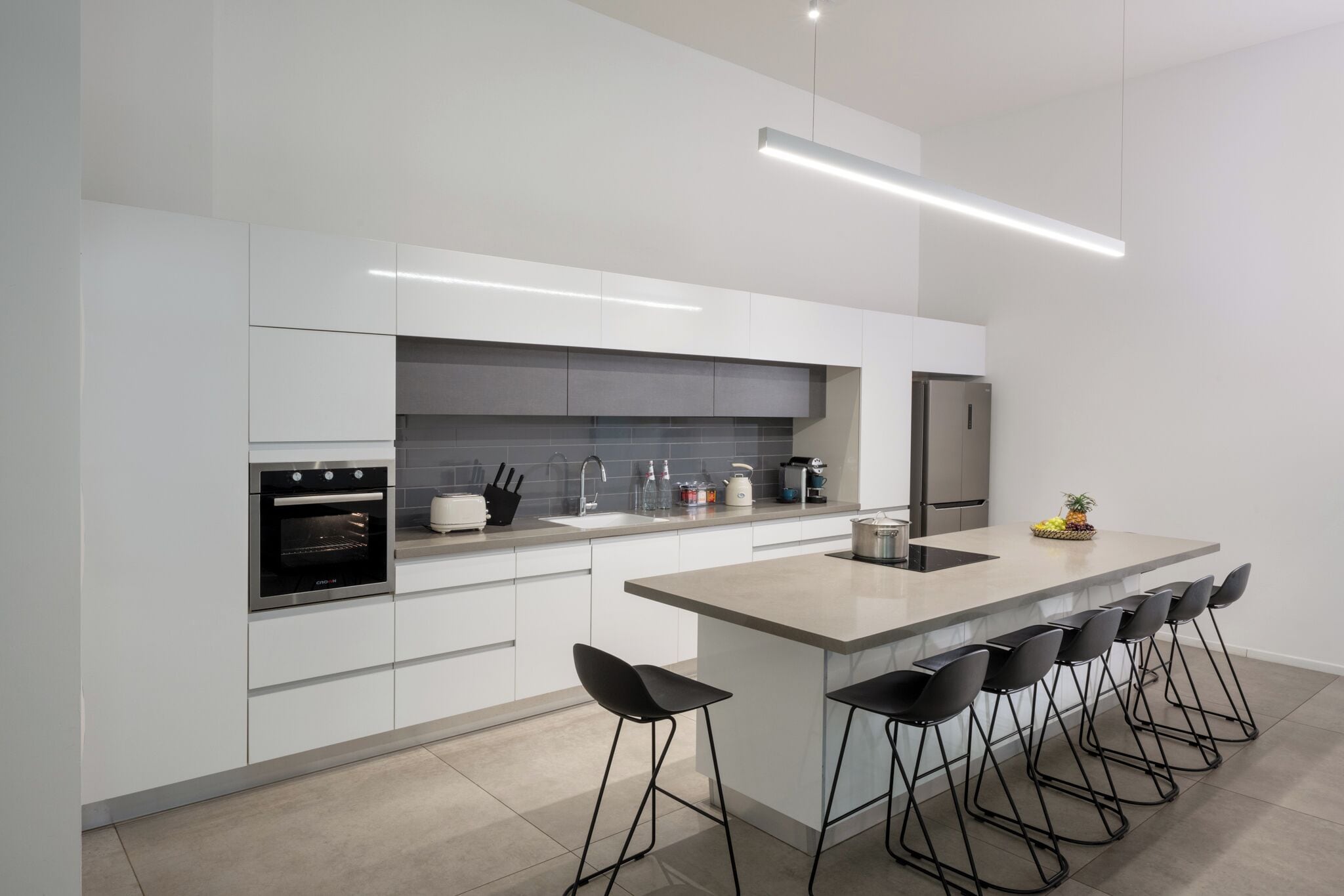 A modern kitchen with white cabinetry, a built-in oven, a stainless steel refrigerator, and a bar area with stools.