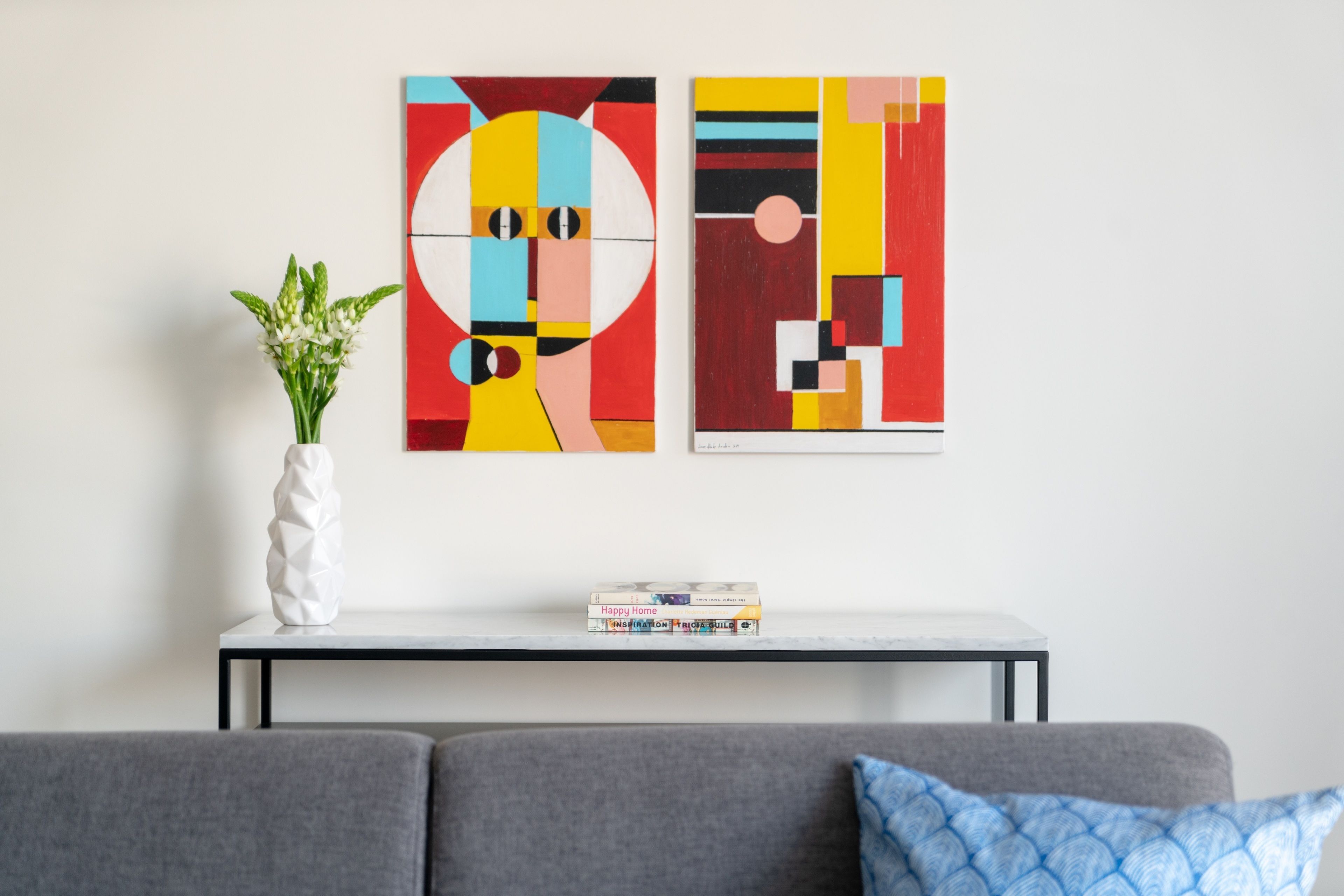 A living room with a gray sofa, a white vase with green plants, and two abstract paintings on the wall.