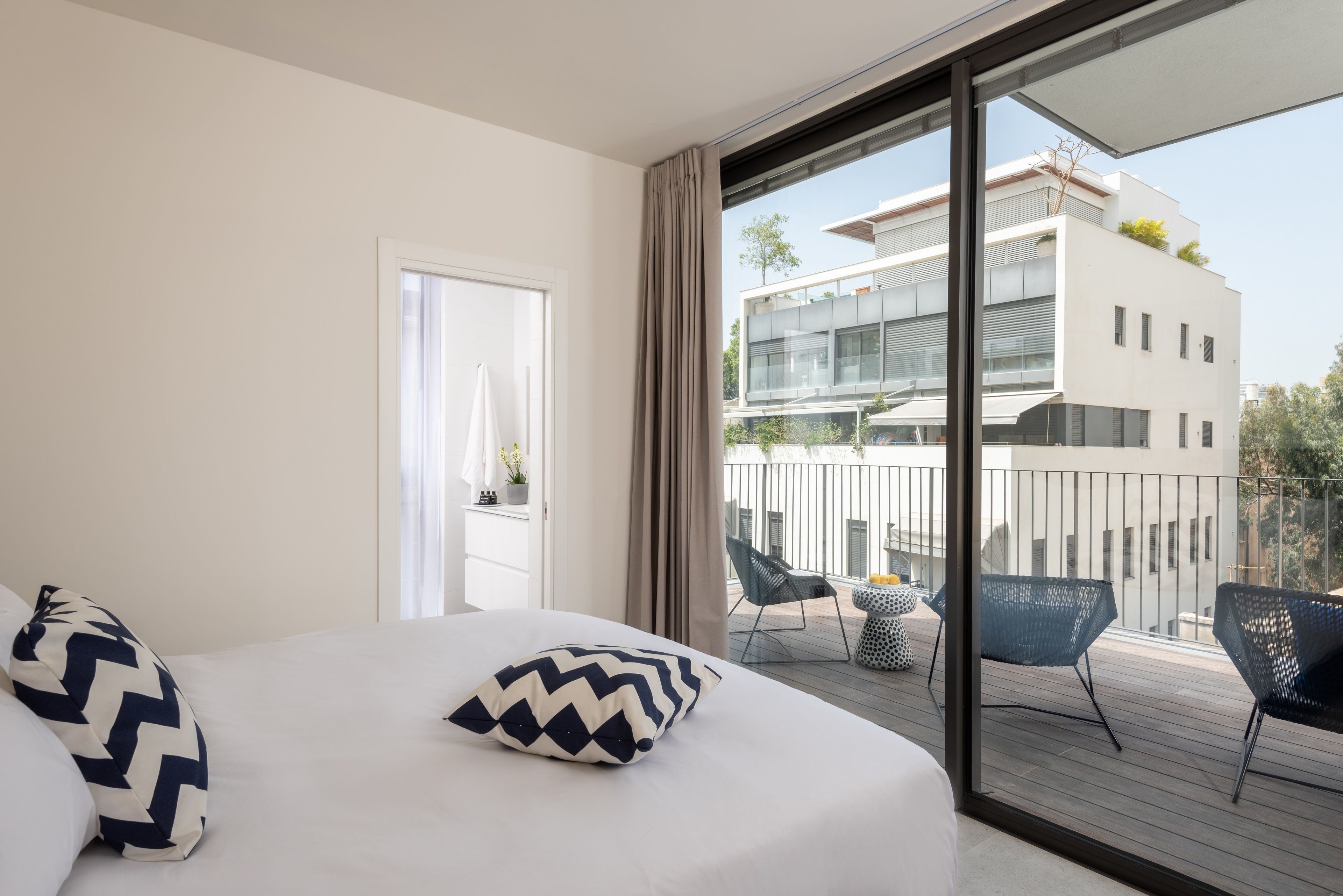 A modern hotel room with a large bed, a balcony with seating, and a view of a white building.