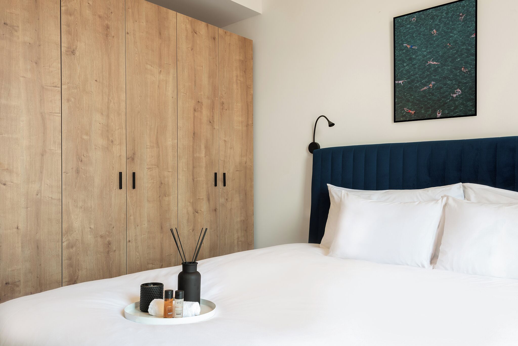 A bedroom with a wooden wardrobe, a bed with white bedding, a blue headboard, and a wall-mounted picture.
