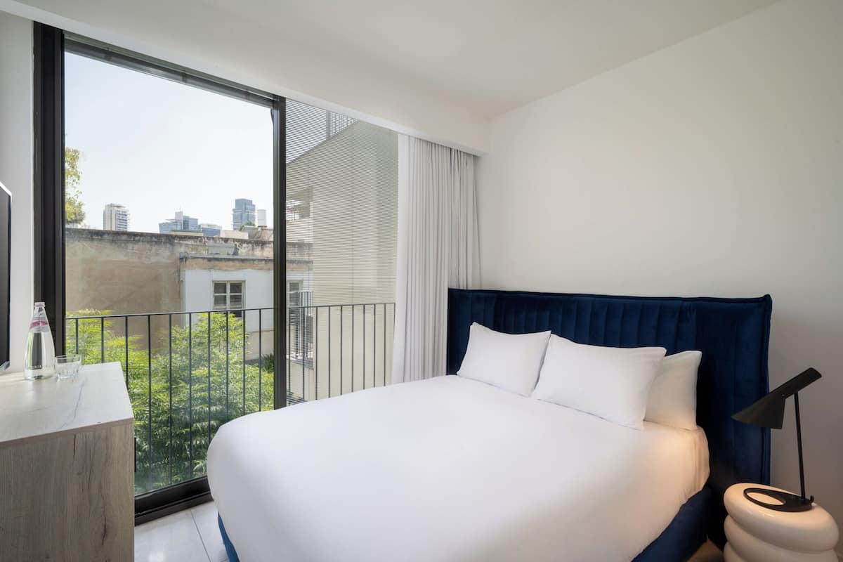Ein Hotelzimmer mit einem großen Bett, einem Nachttisch, einem Schreibtisch und Blick auf die Stadtlandschaft durch ein großes Fenster.