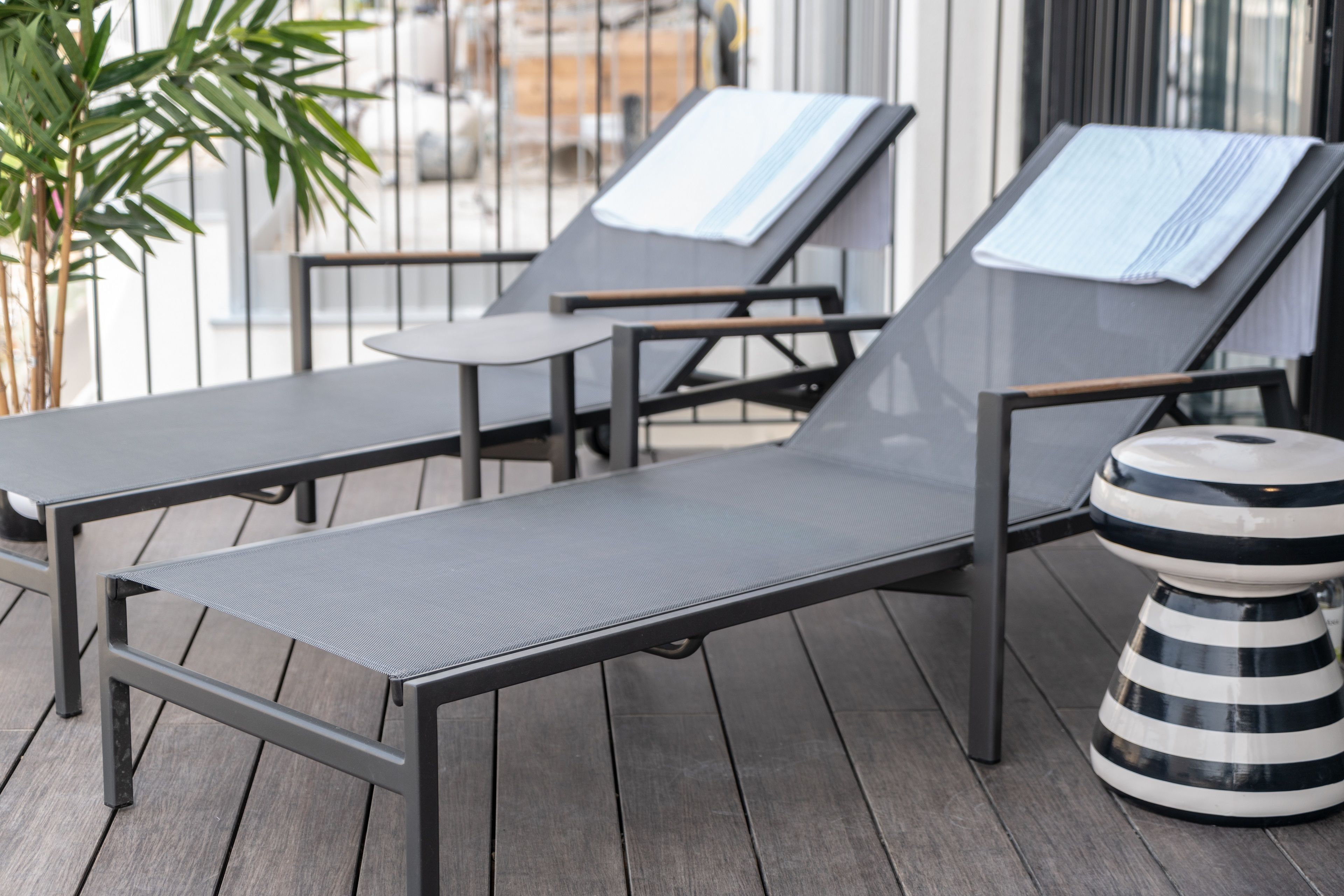 A sun lounger with a metal frame and a cushion, a small table, and a striped stool on a wooden deck.