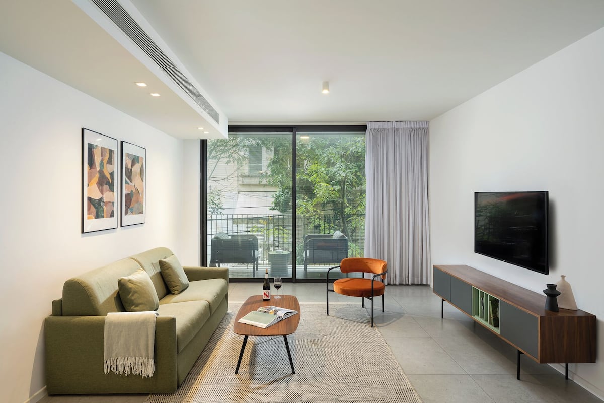 Ein modernes Wohnzimmer mit Sofa, Sessel, Couchtisch, Fernseher und großen Fenstern.