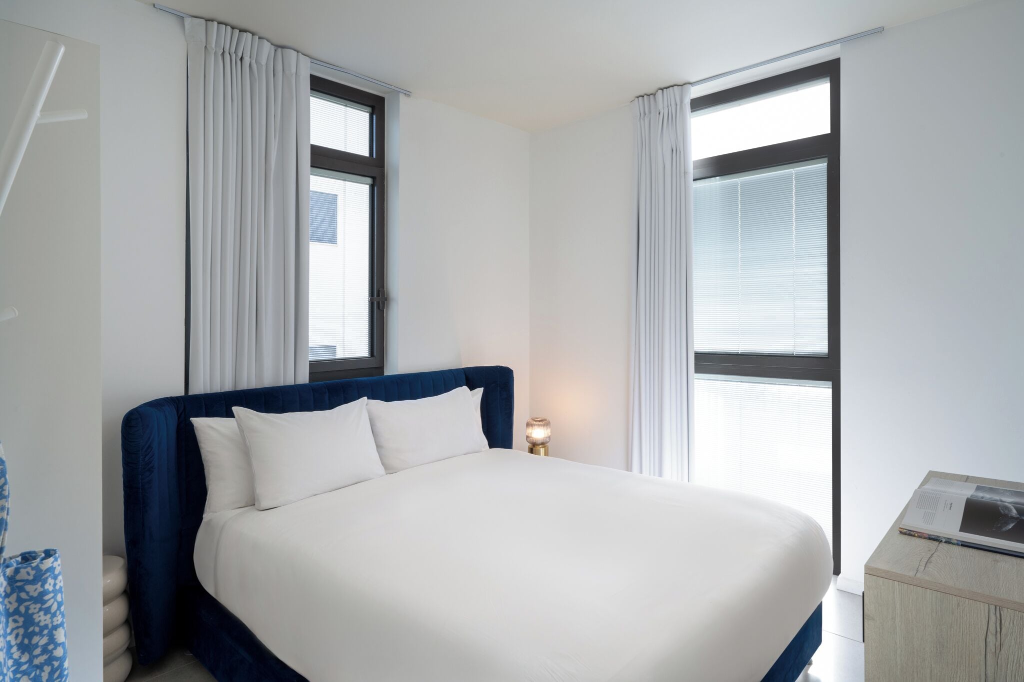 Un lit bien fait, avec du linge de lit blanc, une tête de lit bleue et une table de chevet surmontée d’une lampe.