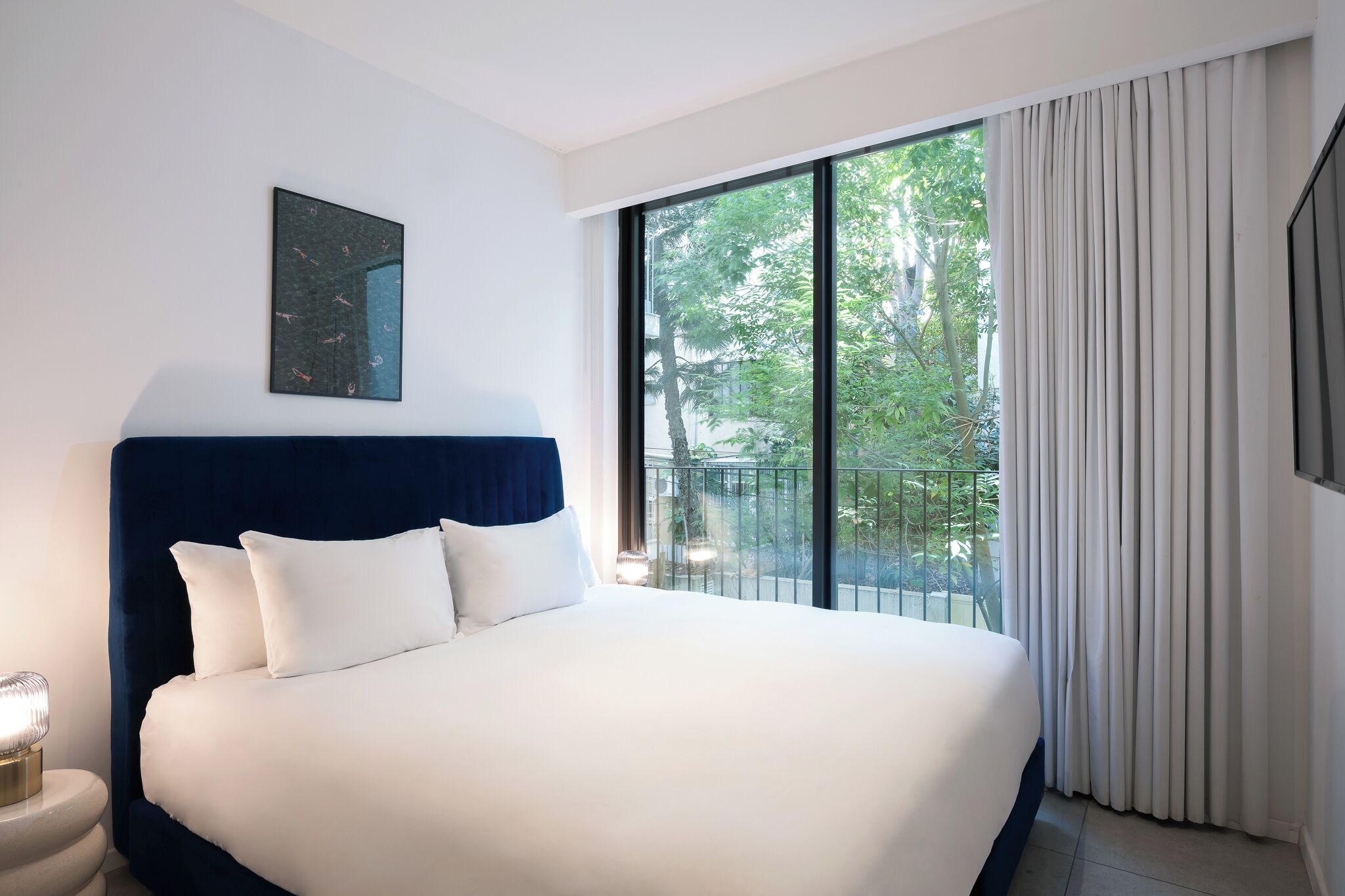 A hotel room with a large bed, a nightstand, a wall-mounted artwork, and a balcony with curtains.