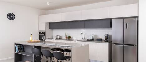 A modern kitchen with a central island, bar stools, a stainless steel refrigerator, and a clock on the wall.