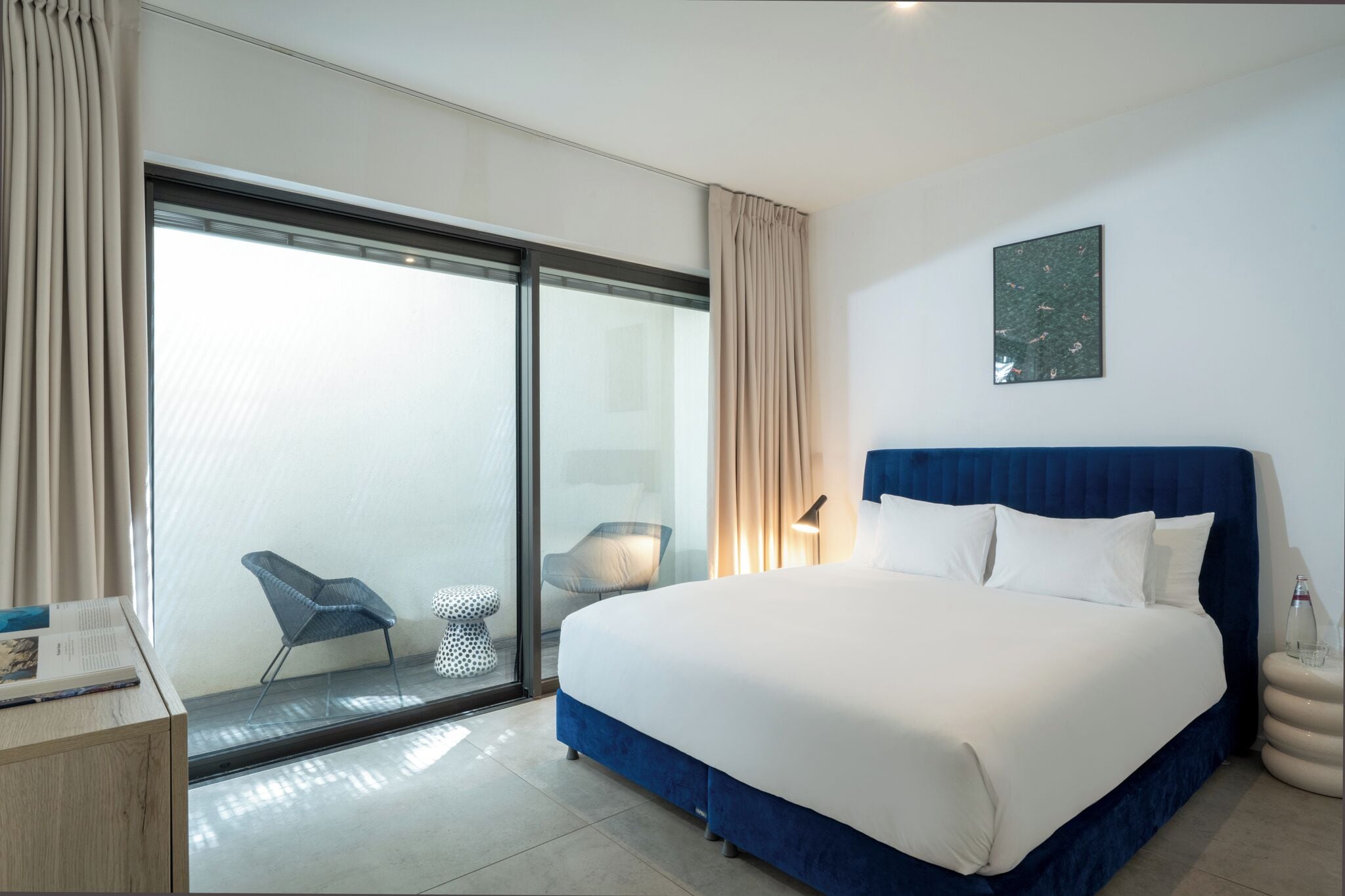 Una habitación de hotel moderna con una cama grande, un vidrio que da a un balcón, una mesita auxiliar pequeña y una silla.