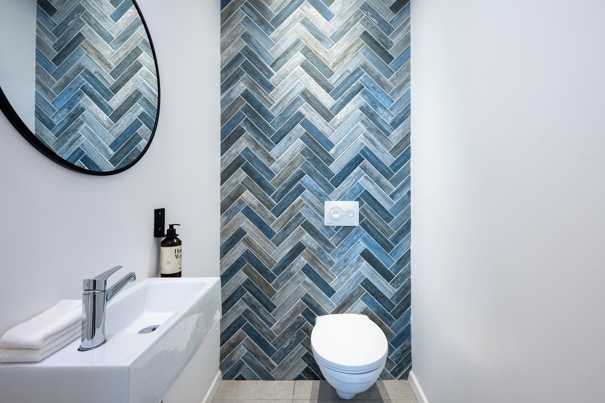 Un baño con pared de diseño espiga, un lavamanos blanco, un grifo cromado, un inodoro y un espejo.