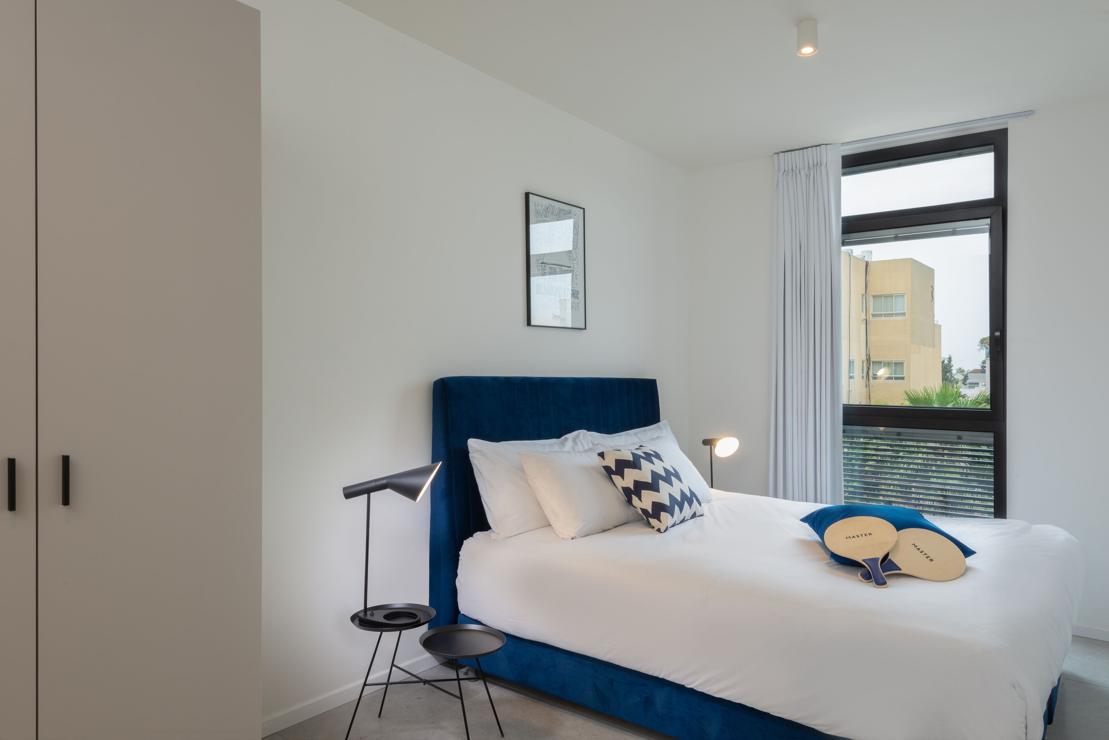 A modern bedroom with a large bed, a floor lamp, and a view of a building through a window.