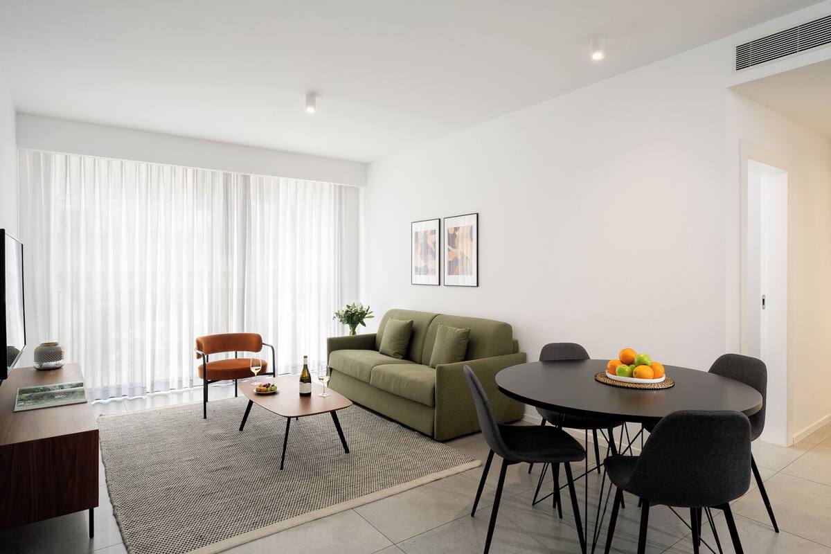 Ein modernes Wohnzimmer mit einer Couch, einem Esstisch mit Stühlen, einem kleinen Couchtisch und einem großen Fenster mit durchsichtigen Vorhängen.