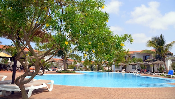 A swimming pool with lounge chairs and umbrellas, surrounded by palm trees and a building with a tiled roof. - Private Self-Catering Apartments (Sal)