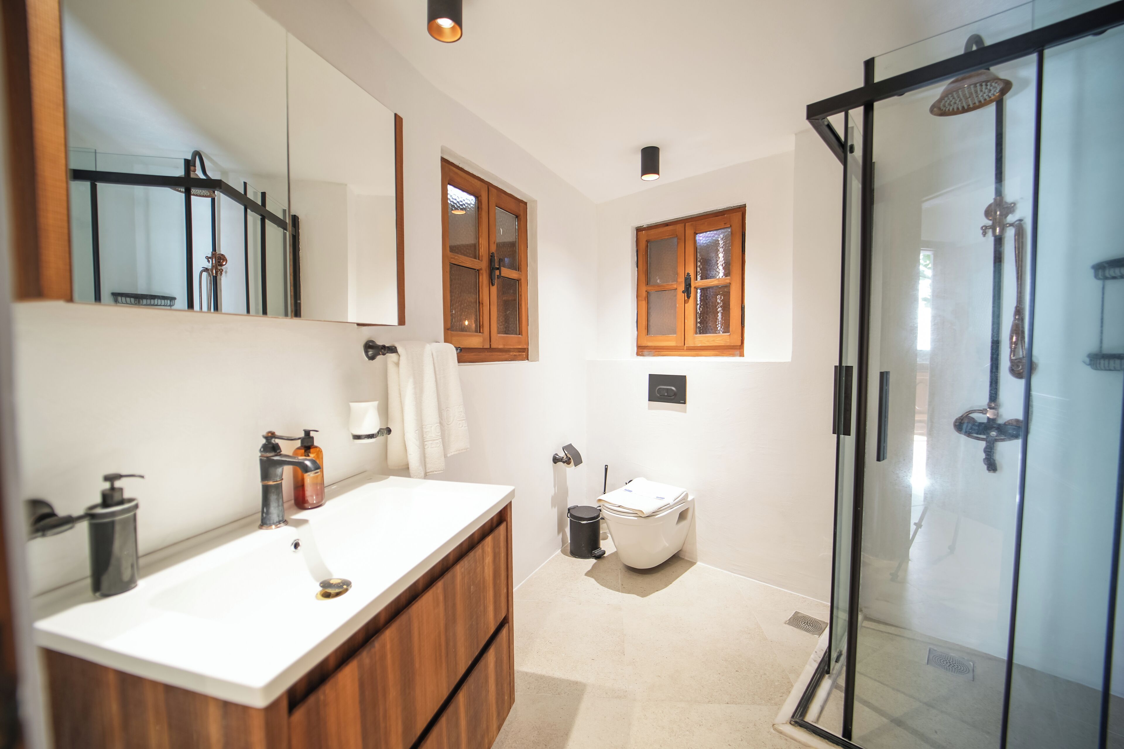 A modern bathroom with a white sink, wooden vanity, a glass shower enclosure, and two wooden-framed windows.