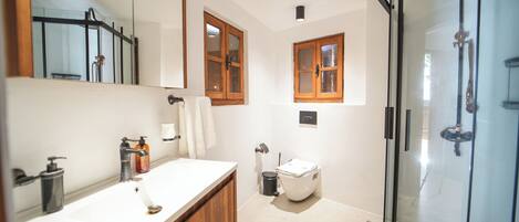A modern bathroom with a white sink, wooden vanity, a glass shower enclosure, and two wooden-framed windows.