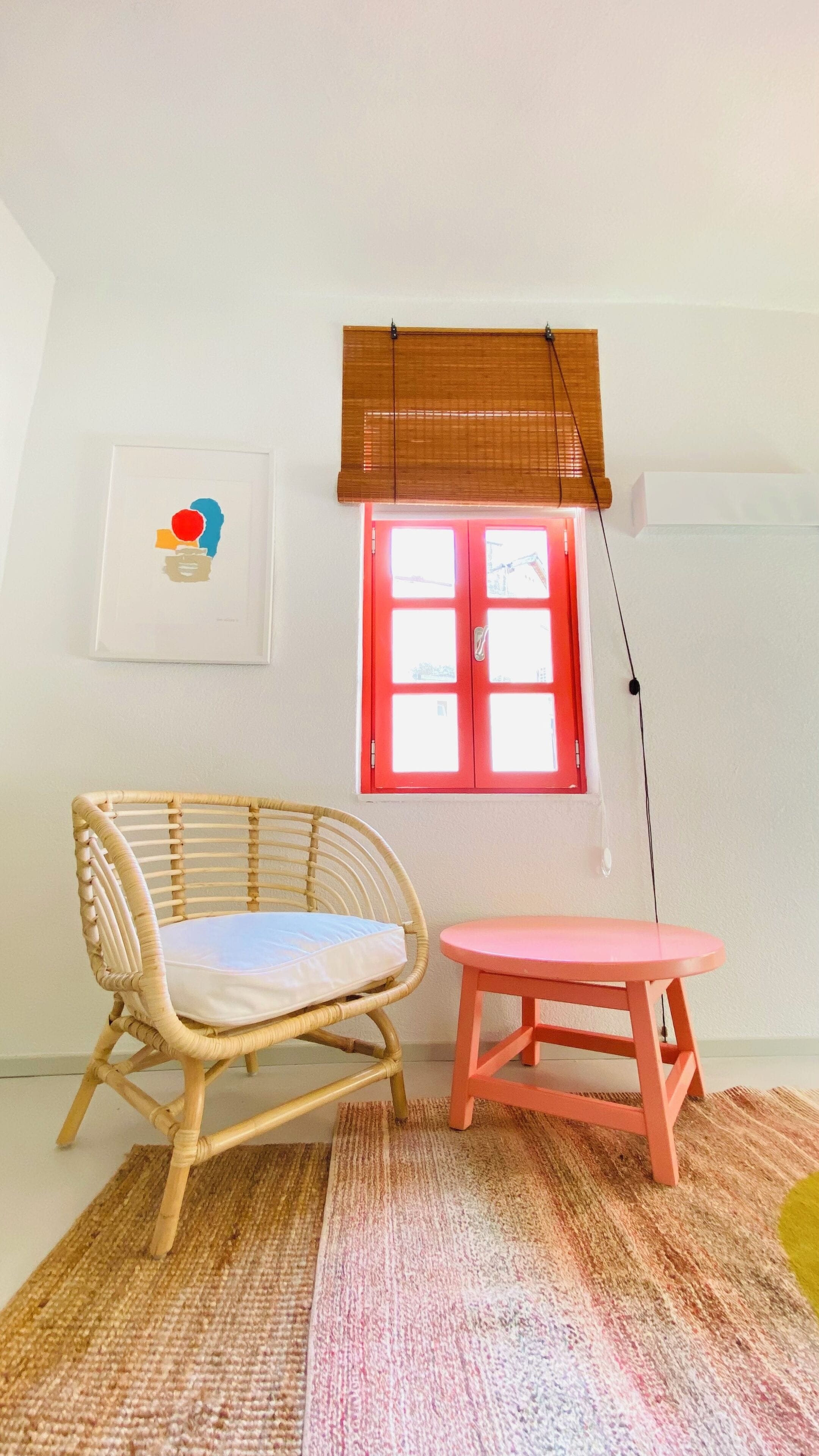 A room with a wicker chair, a small pink table, a red-framed window, and a wall-mounted artwork.