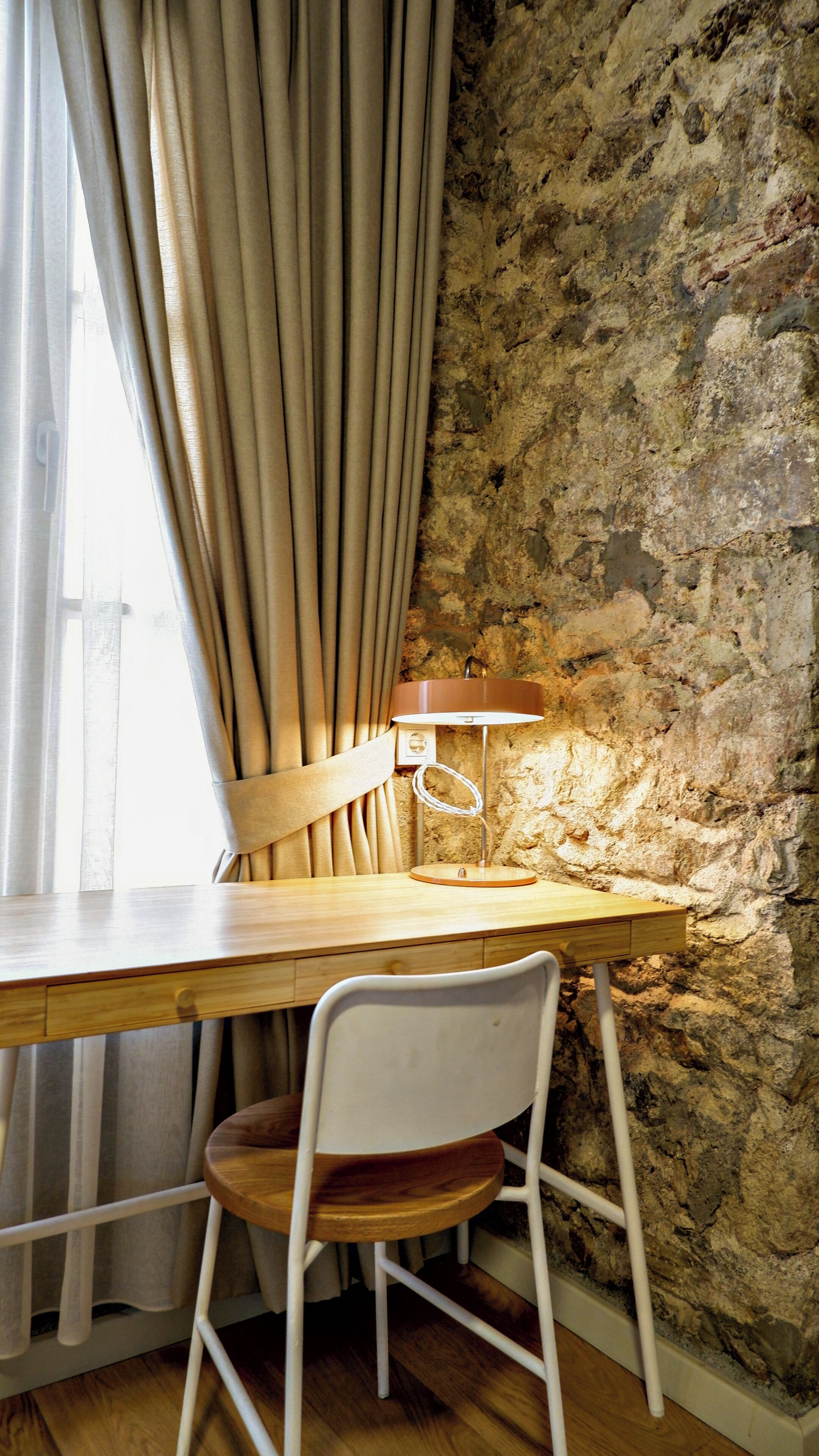 A wooden desk with a chair, a lamp, and a stone wall.