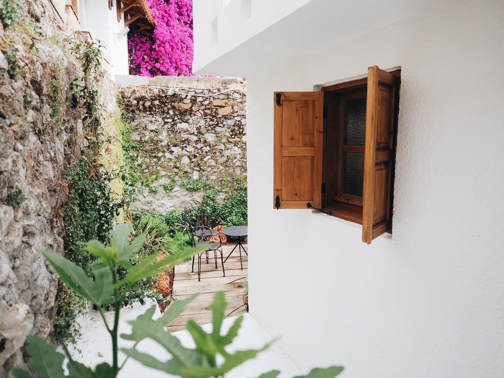 A narrow courtyard with a stone wall, a wooden door, and a small table with chairs.